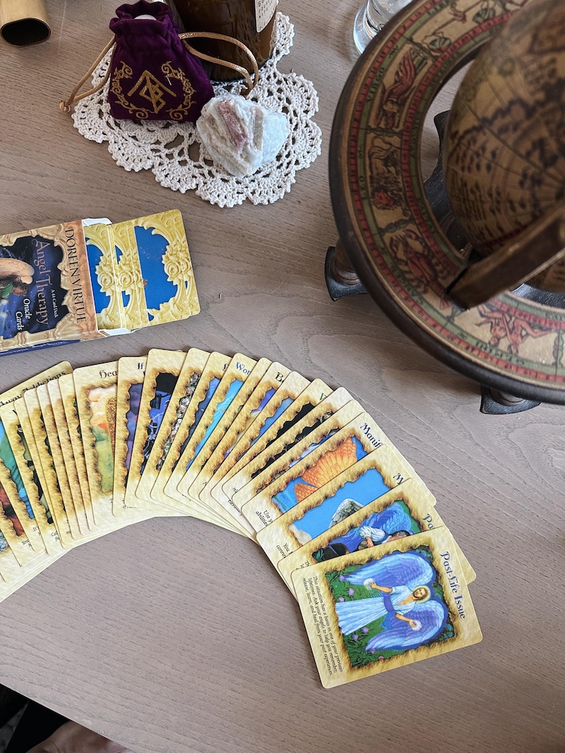 May include: A deck of Doreen Virtue Angel Therapy Oracle Cards fanned out on a light-colored wooden surface. The cards feature colorful illustrations and text. A small purple velvet bag, a white crystal, and a decorative doily are also visible.