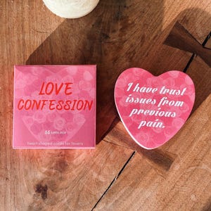 May include: A pink box of "LOVE CONFESSION" cards and a heart-shaped card with the text "I have trust issues from previous pain." The box reads "55 CARD DECK" and "heart shaped cards for lovers."
