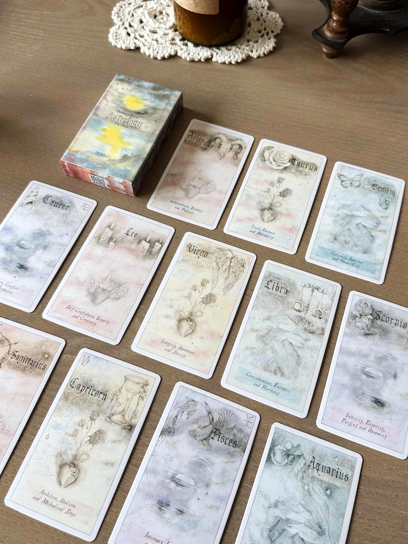 May include: A deck of astrology cards with a watercolor design. The box reads "Astrology" with a sun and moon illustration. Each card features a zodiac sign, with illustrations and text. The cards are laid out on a wooden surface.