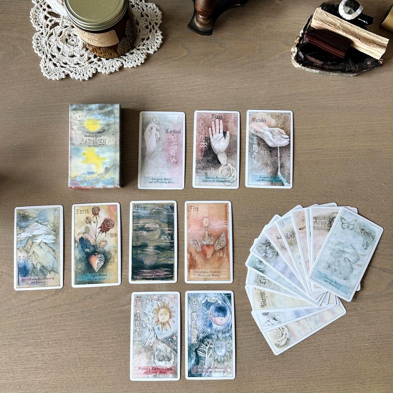 May include: A deck of astrology-themed tarot cards with watercolor illustrations. The cards feature images of hands, celestial bodies, and natural elements. The box reads "Astrology".