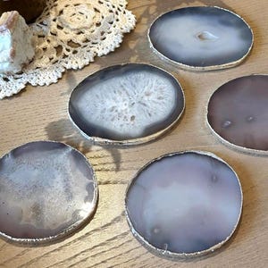 May include: A collection of agate coasters with silver-colored edges. The coasters feature a natural, organic design with shades of gray, white, and purple. The coasters are round and vary slightly in color and pattern.