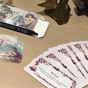May include: A deck of "Sea Melodies" oracle cards with a mermaid illustration on the box. The cards feature nautical-themed artwork and the word "WILD" with the text "Believe in your otherworldly beauty; dance with the waves and drink in the sunshine."