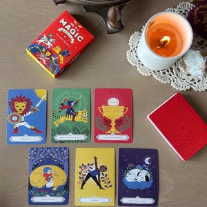 May include: A red box set of "My First Magic Oracle" cards with colorful illustrations. Several cards are displayed, featuring images of a lion, a person on a tiger, a trophy, a person doing yoga, and a sleeping dog. A lit candle and a globe are also in the image.