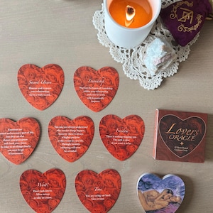 May include: A Lovers Oracle card deck with heart-shaped cards featuring rose patterns and text. A lit candle, a small purple pouch, and a crystal are also present. The deck includes 45 heart-shaped guidance cards.