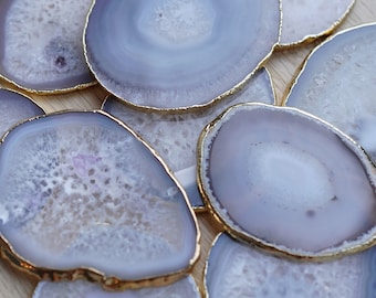 May include: A collection of light purple agate coasters with gold gilded edges. The coasters have a natural, organic shape and a polished finish. The agate slices are arranged on a light wood surface.