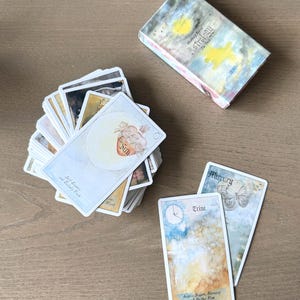 May include: A deck of astrology cards with a box. The cards feature watercolor illustrations and text, including "Sun," "Trine," and "Mercury." The box reads "Humaly Bl Astrology The Deck."