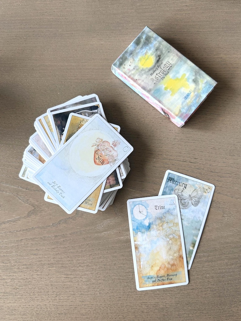 May include: A deck of astrology cards with a box. The cards feature watercolor illustrations and text, including "Sun," "Trine," and "Mercury." The box reads "Humaly Bl Astrology The Deck."