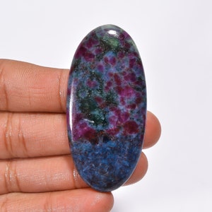 May include: An oval-shaped gemstone with a polished surface. The stone features a blend of colors, including deep red, green, and blue hues. The gemstone is held in a hand, showcasing its size and shape. The stone's pattern is intricate and visually appealing.