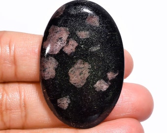Puede incluir: Una gema ovalada con superficie pulida. La piedra es principalmente negra con inclusiones irregulares rosas y verdes. La gema se sostiene en una mano, mostrando su forma. Adecuada para joyería o proyectos de artesanía.