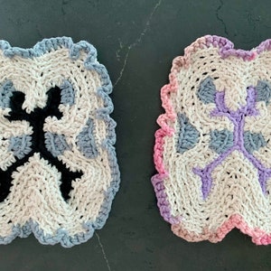 May include: Two crocheted dishcloths with a unique design. One is white with blue and black accents, the other is white with pink and purple accents.
