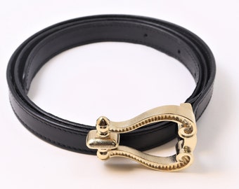 Pode incluir: Um cinto de couro preto com uma fivela de metal em tom dourado polido. A fivela tem um design distinto em forma de U com um pino central vertical e detalhes decorativos em volutas na base, fixada à tira preta lisa com costuras.