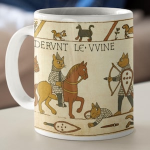 Op de afbeelding: Witte keramische mok met een beige tapijtontwerp met katten in middeleeuwse harnassen, paarden berijden en pijl en boog gebruiken. De mok heeft de tekst "DERVNT LE VVINE" en een wit handvat.