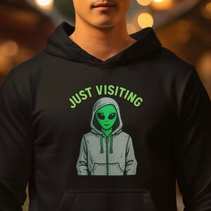 Puede incluir: Sudadera con capucha negra con bolsillo delantero y capucha con cordón. El gráfico en la parte delantera presenta un extraterrestre verde con el texto "JUST VISITING" en verde arriba.