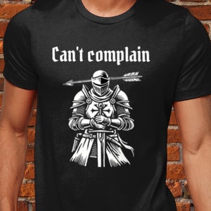 Peut inclure: T-shirt noir avec une illustration blanche d'un chevalier avec une flèche dans son casque. Le texte "Can't complain" est au-dessus du chevalier.