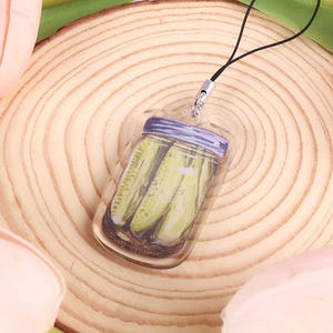 May include: A clear acrylic charm depicting a jar of pickles with a purple lid, attached to a black lanyard. The background features a wooden slice and pink and yellow tulips.