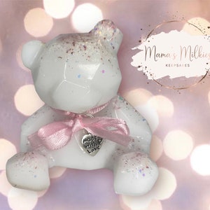 Op de afbeelding: Witte geometrische teddybeer van hars met een roze lint en een zilveren bedel met de tekst "Made with Love". De beer zit op een roze en witte glinsterende achtergrond. Mama's Milkies Keepsakes.