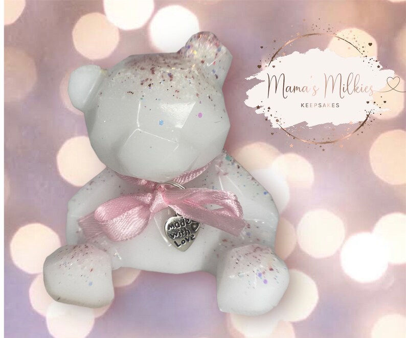 Op de afbeelding: Witte geometrische teddybeer van hars met een roze lint en een zilveren bedel met de tekst "Made with Love". De beer zit op een roze en witte glinsterende achtergrond. Mama's Milkies Keepsakes.