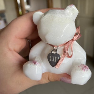 Op de afbeelding: Witte glitterhars berenfiguur met een roze lint en een zilveren hartbedel met de tekst "HELP".