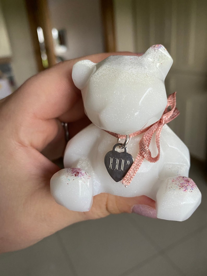 Op de afbeelding: Witte glitterhars berenfiguur met een roze lint en een zilveren hartbedel met de tekst "HELP".