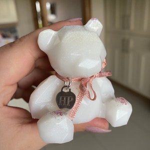 Op de afbeelding: Witte glinsterende geometrische berenfiguur met een roze lint en een zilveren bedel met de tekst "HIP".