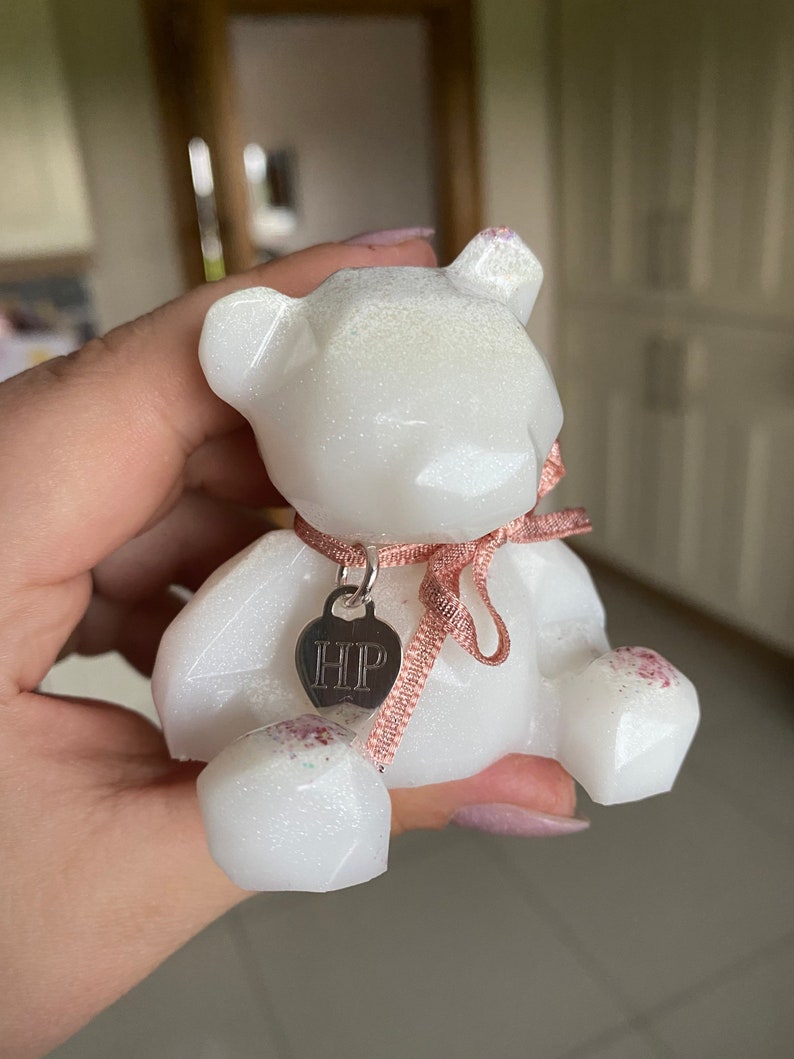 Op de afbeelding: Witte glinsterende geometrische berenfiguur met een roze lint en een zilveren bedel met de tekst "HIP".