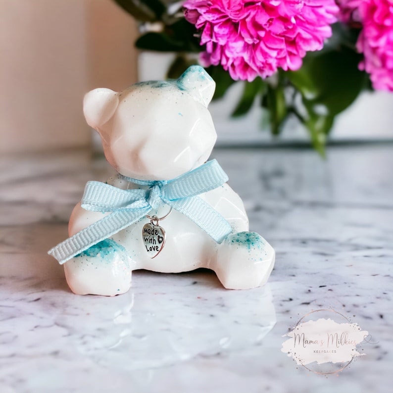 Op de afbeelding: Witte keramische teddybeer met een blauw lint en een zilveren bedel met de tekst "made with love".