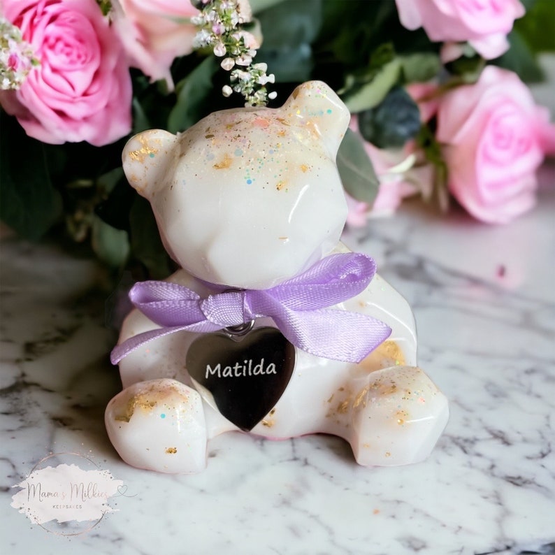 Op de afbeelding: Witte keramische berenfiguur met een paars lint en een zilveren hartbedel met de naam Matilda erop gegraveerd. De beer zit op een wit marmeren oppervlak.