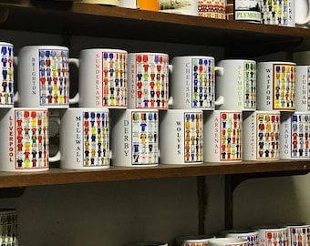 May include: White ceramic mugs with various soccer team jersey designs, including Cardiff, Liverpool, and Arsenal, are displayed on wooden shelves. The mugs are arranged in rows, showcasing a variety of team colors and patterns. The mugs are a standard size.