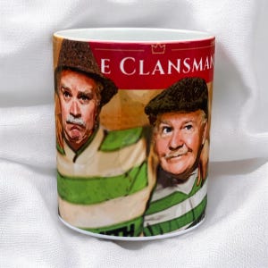 May include: A white ceramic mug with a green and white striped design. The mug features a photo of two men wearing green and white striped jerseys. The text "The Clansman" is printed on the mug.
