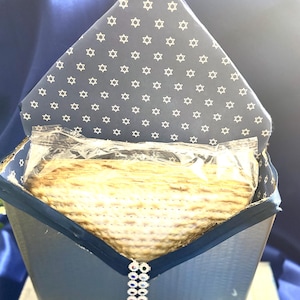 May include: A blue box with a white star pattern on the lid, containing a stack of matzah wrapped in plastic. The box is decorated with a blue ribbon and a string of rhinestones.