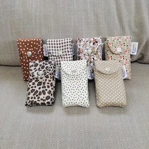 Könnte beinhalten: Acht kleine Stoffbeutel mit verschiedenen Mustern, darunter braune Sterne, braunes und weißes Gingham, Blumen, Punkte und Leopardenmuster. Jeder Beutel hat einen Knopfverschluss und ein kleines Etikett mit der Aufschrift "Mit Liebe gemacht".