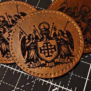 Peut inclure: Écussons ronds en cuir marron avec un motif graphique noir. Le motif représente des armoiries avec une croix, des anges et du texte. Les écussons sont cousus sur les bords et sont sur une surface quadrillée noire.