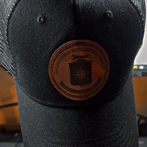 Op de afbeelding: Zwarte baseballcap met een mesh achterkant en een ronde leren patch. De patch heeft de tekst "ENGINEERING INTELLIGENCE AGENCY" en een schildontwerp. Klassieke stijl, geschikt voor casual wear.