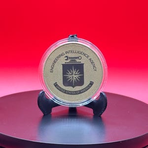 May include: A gold-colored coin with the text "ENGINEERING INTELLIGENCE AGENCY" and "UNITED STATES OF AMERICA" is displayed on a black stand. The coin features a shield design with a starburst and tools.