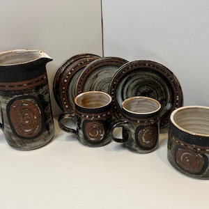 May include: A ceramic set consisting of a pitcher, two mugs, a bowl, and three saucers. Each piece features a dark brown and grey mottled glaze with a prominent rust-colored spiral design, a dark band at the rim, and small white dot accents.