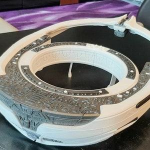 May include: A white and gray 3D printed model of a futuristic spaceship. The model is detailed with intricate patterns and textures. The spaceship has a circular design with a central opening.