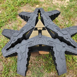May include: A black and gray 3D printed model of a spaceship with a pyramid-shaped structure in the center. The model is sitting on a bed of green grass.