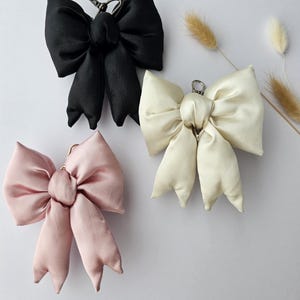 May include: Three satin bow keychains in black, cream, and blush pink. Each bow has a metal clasp for attaching to keys or bags. The bows are arranged on a white surface with dried floral accents.