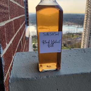 Puede incluir: Una botella transparente cuadrada con una tapa azul oscuro, llena de un líquido marrón dorado. Una etiqueta blanca dice "Truths Told Tarot Black Walnut Bath". La botella está colocada contra un muro de ladrillos y un paisaje urbano.