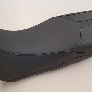 May include: A black motorcycle seat with a contoured design. The seat has a smooth, leather-like texture and features stitched detailing along the edges. The seat's shape is designed for comfort and support during rides.