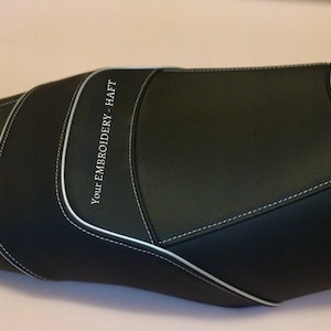 May include: Black motorcycle seat with white stitching and the text "Your Embroidery - Haft" embroidered on the side.