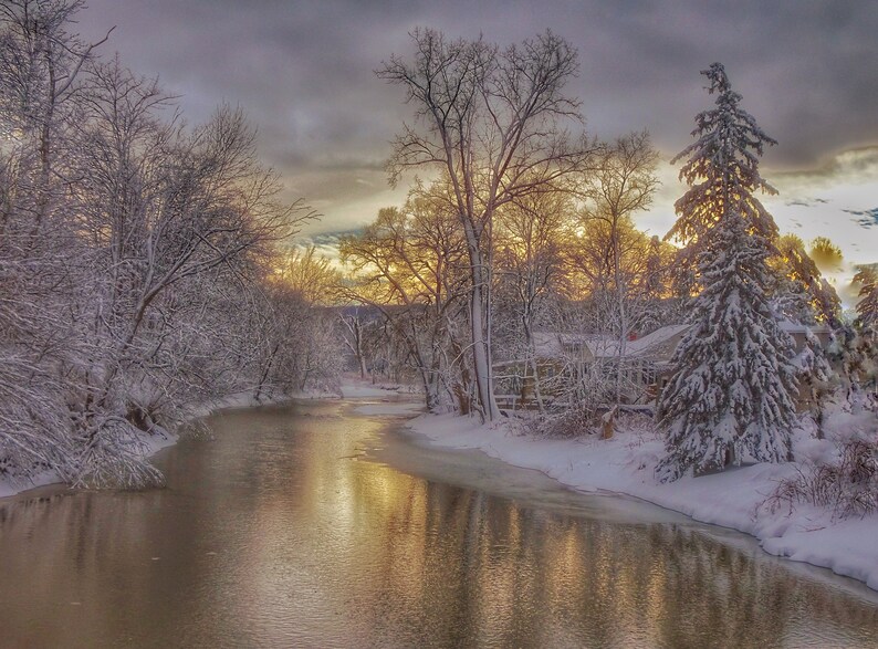 May include: A snowy winter scene with a river flowing through a forest. The trees are covered in snow and the river is reflecting the golden light of the setting sun.