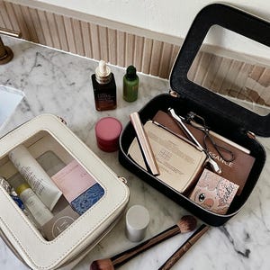 May include: Two open cosmetic bags, one cream and one black, filled with makeup and skincare products. The cream bag has a clear window, and the black bag is open to reveal various beauty items. Makeup brushes and skincare bottles are also visible.