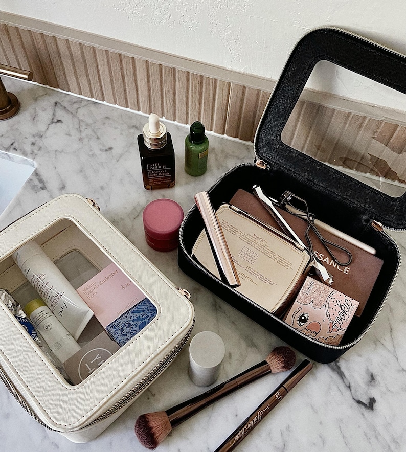 May include: Two open cosmetic bags, one cream and one black, filled with makeup and skincare products. The cream bag has a clear window, and the black bag is open to reveal various beauty items. Makeup brushes and skincare bottles are also visible.