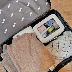 May include: An open black suitcase filled with travel items. A beige sweater, a white cosmetic bag containing toiletries, and a patterned fabric bag are visible. The suitcase lining is grey with the word "AWAY" printed repeatedly.