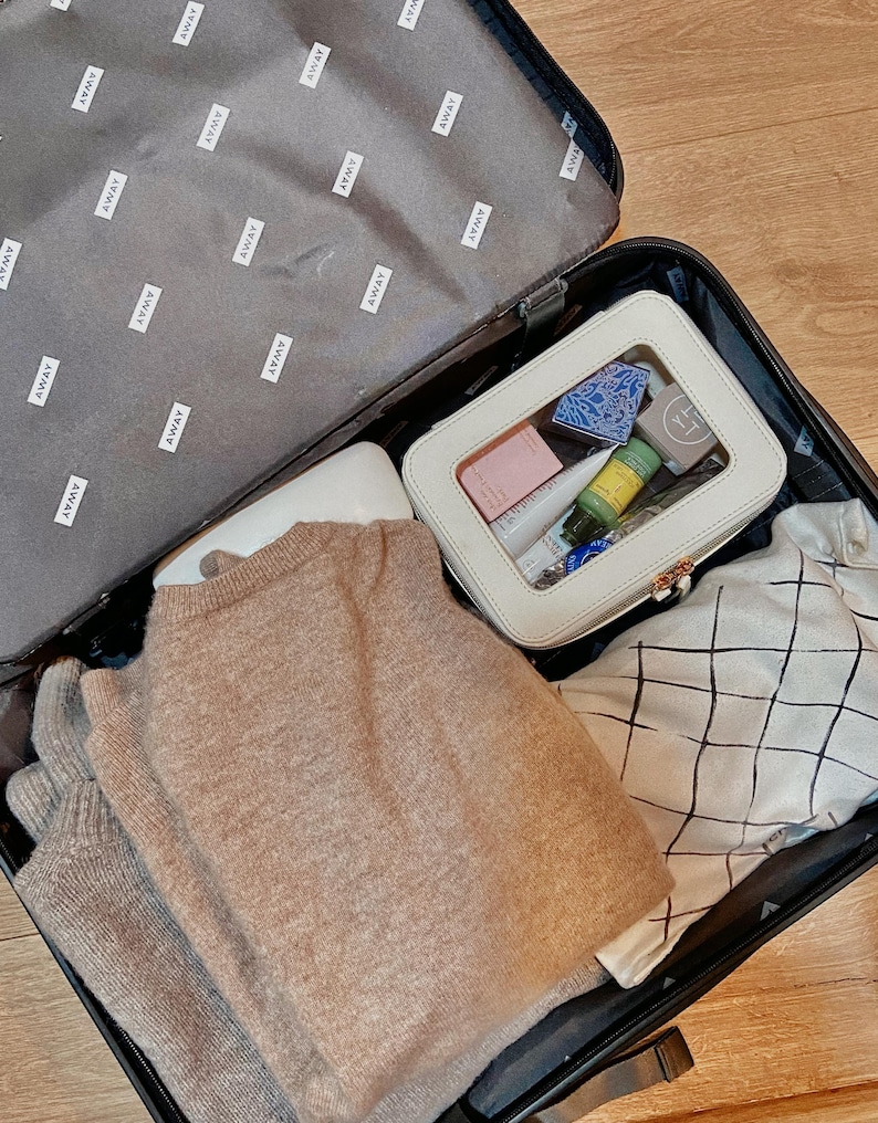 May include: An open black suitcase filled with travel items. A beige sweater, a white cosmetic bag containing toiletries, and a patterned fabric bag are visible. The suitcase lining is grey with the word "AWAY" printed repeatedly.