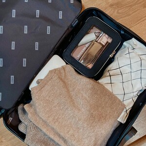 May include: A black suitcase, open and packed with travel items. Inside is a black cosmetic bag with "MODERN REN" text, a beige sweater, a white item, and a patterned shirt. The suitcase interior is black with "AWAY" branding.