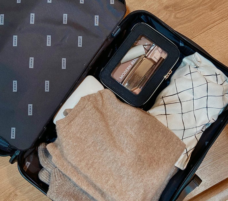 May include: A black suitcase, open and packed with travel items. Inside is a black cosmetic bag with "MODERN REN" text, a beige sweater, a white item, and a patterned shirt. The suitcase interior is black with "AWAY" branding.