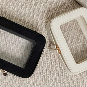 May include: Two rectangular cosmetic bags, one black and one cream, each with a clear window. The bags have a zip closure and gold-coloured hardware. They are on a textured, neutral-coloured surface.
