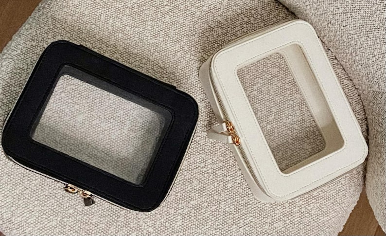 May include: Two rectangular cosmetic bags, one black and one cream, each with a clear window. The bags have a zip closure and gold-coloured hardware. They are on a textured, neutral-coloured surface.